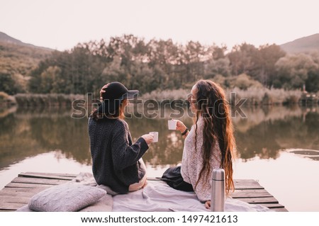 Two female friends in knitted warm sweaters having picnic near lake with autumn forest and lake on the background. Cozy fall atmosphere.