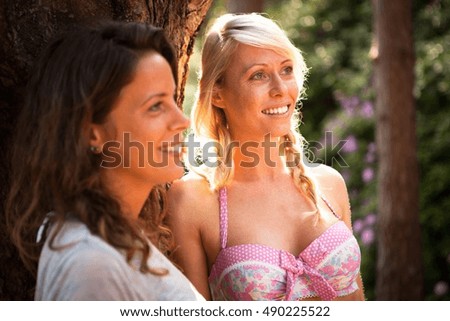 Two female friends leaning against tree in forest