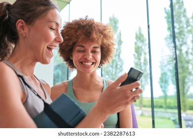 Two Female Friends Look At Mobile Phone Wearing Gym Clothing At Gym Or Yoga Studio For Exercise - Powered by Shutterstock