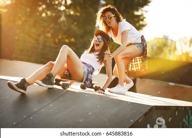Two female friends playing with skateboard at the skate park.One girl pushing other from behind.Laughing and fun.