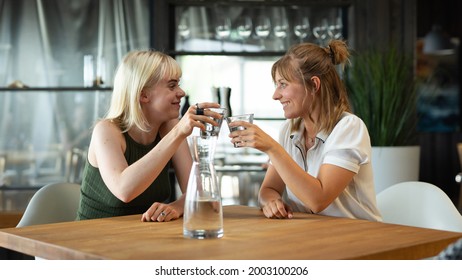 Two Female Friends Raising A Glass While Feeling Happy And Enjoying Eachothers Company In A Modern Restaurant Sitting Behind A Wooden Table With Glasses Of Water. 