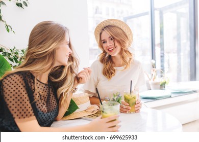 Two Female Friends Resting Together In Favorite Cafe And Sharing News Happy To See Each Other. Indoor Portrait Of Amazing Girls In Cute Dresses Drinking Cocktails And Talking About Something Funny.