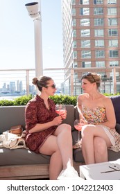 Two female friends sitting in the sun on a trendy rooftop bar in Manhattan. Two girls having a drink, relaxing at a rooftop bar on the weekend in the city. Having fun drinking on a rooftop in NYC.
