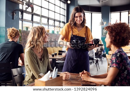 Two Female Friends Sitting At Table In Coffee Shop Being Served By Waitress