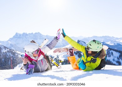 two female friends with ski and snowboard equipment having fun and high five with snow in the air.