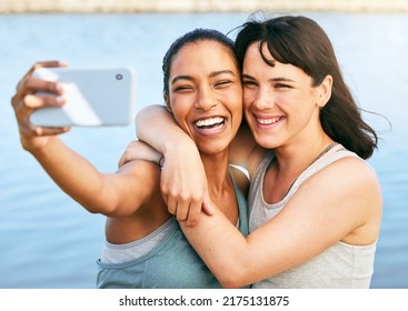 Two Female Friends Taking Selfies After A Workout In Nature And Standing In Front Of A Lake Using A Smartphone. Young Latino Female Using Her Phone To Take A Selfie Of Herself And Her Friend