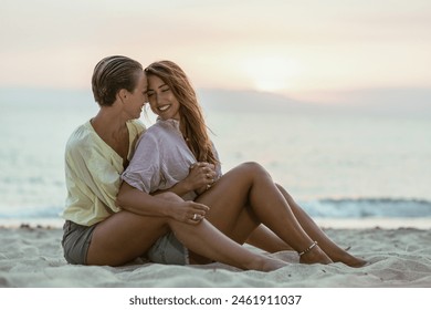 A two female girl friends are embracing each other while sitting on a sandy beach, with the sea in the background. - Powered by Shutterstock