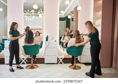 Two female hairstylists curling client hair and doing hairstyle in modern beauty salon. Young women sitting in chairs in front of mirrors while hairdressers styling clients hair. - Powered by Shutterstock