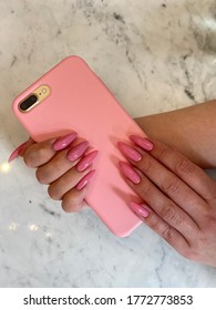 Two Female Hands With Long Pink Nails Hold Pink Phone On Marble Background.