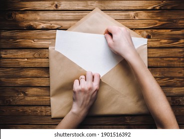 Two Female Hands Open A Large Brown Envelope And Take Out A Letter From It. Wooden Background, Copy Space And Mockup.