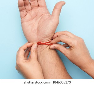 Two Female Hands Tie A Red Woolen Thread On The Wrist Of A Male Hand, Blue Background, Top View