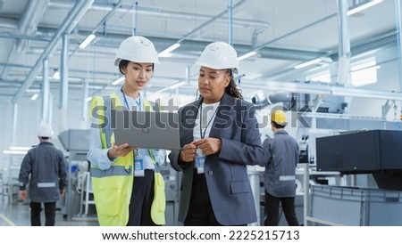 Two Female Heavy Industry Employees in Hard Hats at Factory. Discussing Job Assignments at Industrial Machine Facility, Using Laptop Computer. Asian Engineer and African American Technician at Work.