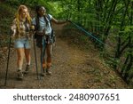 Two female hikers walking down a forest hiking trail wearing shorts and hiking boots with one of them pointing at something with her trekking pole.