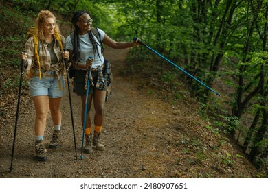 Two female hikers walking down a forest hiking trail wearing shorts and hiking boots with one of them pointing at something with her trekking pole.