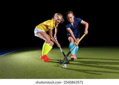 Two female hockey players focus intensely as they battle for the ball on a well-lit field. One wears a yellow jersey and brightly colored socks, while the other sports a dark blue uniform. - Powered by Shutterstock