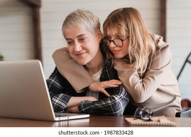Two female LGBT lesbian couple best girlfriends students hugging embracing watching movie webinar on laptop studying e-learning together, doing paperwork, paying bills online in home office
