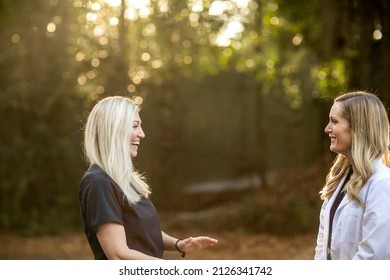 Two Female Medical Professionals In Black Scrubs And One Is Wearing A White Lab Coat Standing Outside Discussing A Case.