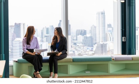 Two female office workers using a laptop to discuss for a new business project in an office with cityscape background. Informal discussion between two young staff with urban landscape background