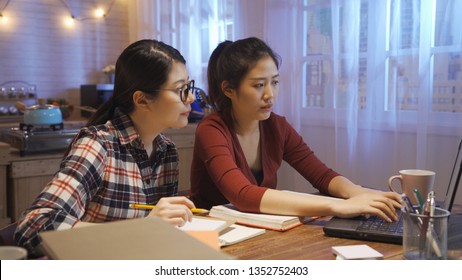 Two Female People Doing Group Study At Wooden Table With Girl Typing Keyboard On Laptop. University Students Researching Information Project On Computer Discussing Looking At Screen In Night Home.