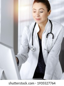 Two female physicians are discussing medical therapy, while standing at the table in a sunny clinic office. Doctors use pc computer at work. Teamwork in medicine