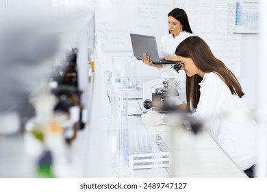 Two female scientists in a bright laboratory conducting research one is using a microscope while the other is working on a laptop with lab equipment around. - Powered by Shutterstock