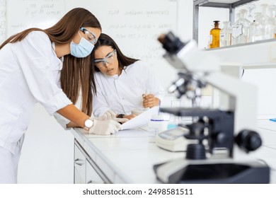 Two female scientists in protective gear working in a laboratory, analyzing documents, and using scientific equipment to conduct experiments and research. - Powered by Shutterstock