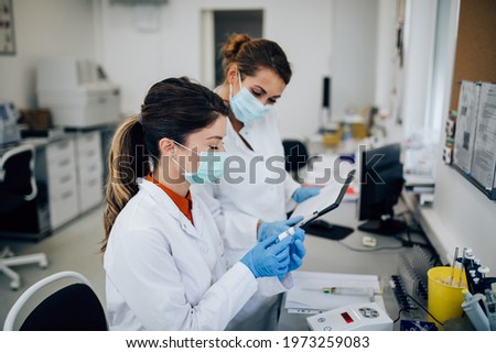 Two female scientists or technicians with face protective masks work in laboratory on human blood samples.	