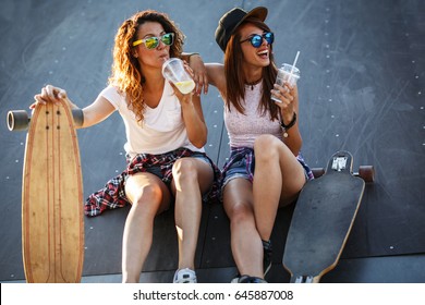 Two Female Skaters Friends Sitting On Ramp At The Skate Park And Drinking Juice .Laughing And Fun.