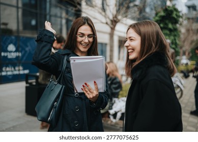 Two female students review notes and laugh while standing outside on campus. The friends study together outdoors in an urban setting, sharing papers and conversation.