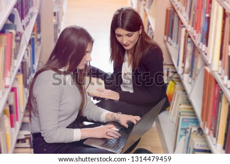 Two Female students taking notes from a book at library, using  laptop and sitting on the floor with a book in the library between the bookshelf and doing assignments in college library.