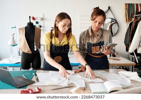 Two female tailors choosing sewing paper patterns by workplace in studio