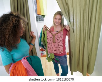 Two Female Teenagers In The Trial Room Holding A Bunch Of Outfits To Try.