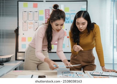 Two female uiux designers collaborating and brainstorming while reviewing a mobile app interface on paper in a modern office. Planning and strategizing development - Powered by Shutterstock