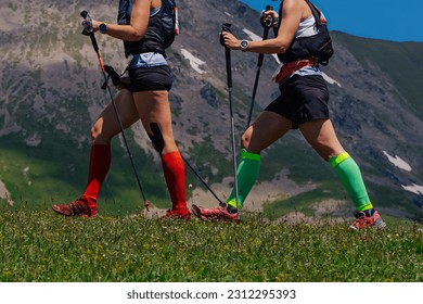 two female walk trail on green meadow in background mountain, compression socks on his feet and trekking poles in hands