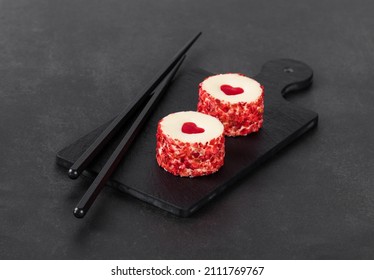 Two festive creamy white chocolate mousse cakes with a jelly heart, in the form of round sweet sushi, sprinkled with small pieces of freeze-dried strawberries on a serving board on a dark background