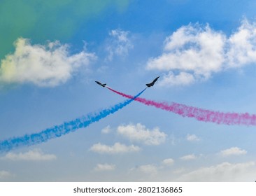 Two fighter jets perform a dynamic crossover maneuver in blue sky, leaving red and blue smoke trails against white clouds in an aerial display.