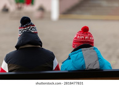 Two figures sit side by side on a bench, wearing winter clothes and specific hats. They watch a winter scene while the weather is cold.
