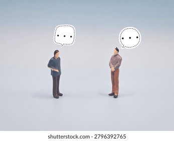 Two figurines with speech bubbles pause in dialogue, representing communication, hesitation, silence, contemplation, and interpersonal connection in abstract human exchange.