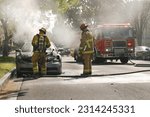 Two fire fighters in full protective gear examine the remains of a partially melted car after they put the engine fire out. A firetruck, fire hoses, and police motorcycle are in the background.