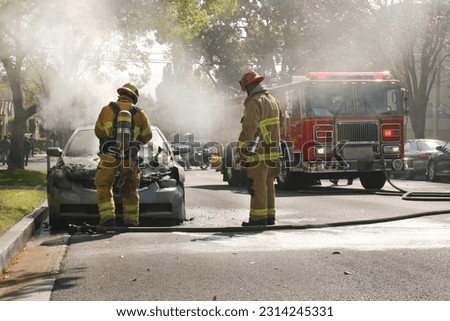 Two fire fighters in full protective gear examine the remains of a partially melted car after they put the engine fire out. A firetruck, fire hoses, and police motorcycle are in the background.