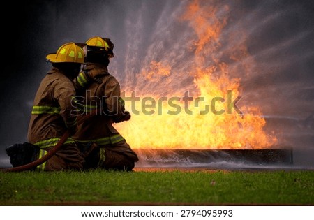 Two firefighters in protective gear work together to extinguish a large, roaring fire, demonstrating teamwork and courage in a high-risk situation