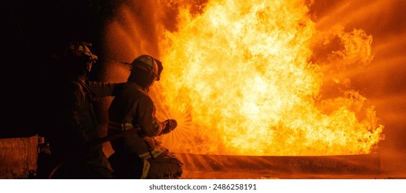 Two firefighters are standing in front of a fire, one of them is holding a hose. The fire is very large and the firefighters are trying to put it out