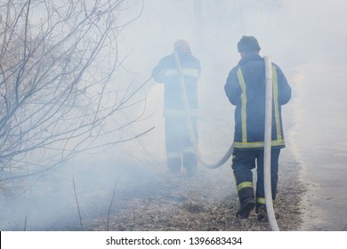 Two Fireman In Smoke Battle Forest Fire. Brave Firefighter Goes Into Smoke To Put Down A Wild Fire, Other Fireman Holds And Carry Hose