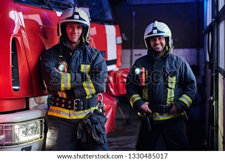 Two firemen wearing protective uniform standing next to a fire engine in a garage of a fire department, smiling and looking at a camera