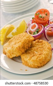 Two Fish Cakes On A Plate With Salad And Sliced Lemons