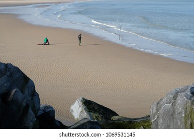 Two fishermen on an empty beach, with rocks in the foreground