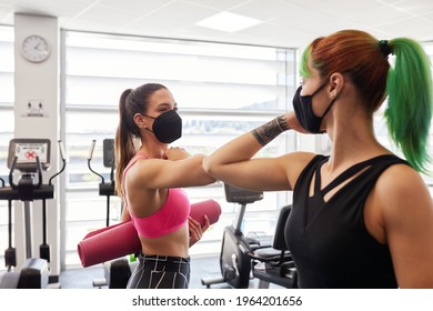 Two fit woman friends greeting in gym by tapping elbows to keep social distancing rules during Coronavirus disease pandemic. Wearing face masks and holding yoga mats - Powered by Shutterstock