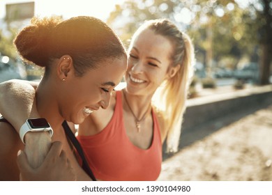 Two Fit Young Women In Sportswear Laughing While Standing Arm In Arm Outside On A Sunny Day After A Workout Together