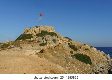 Two flags flutter at the peak of a rocky hill by the calm sea, illuminated by bright sunlight on a summer day. - Powered by Shutterstock