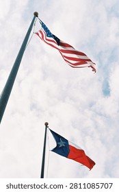 Two flags fly simultaneously on separate tall flagpoles against a partly cloudy sky, photographed from a dramatic low upward angle. The American flag dominates the upper portion of the frame, its red, white, and blue stripes and star field rippling fully extended in the wind. Below and behind it on a second pole, the Texas state flag — with its bold vertical blue stripe, lone star, and red and white horizontal bands — waves in matching fashion. The low perspective and cloud-filled sky give the image a sense of patriotic grandeur and civic pride. The film capture adds a slightly warm, vintage quality to the colors that keeps the image from feeling overly produced. Widely applicable for political, civic, governmental, travel, and Americana editorial contexts.